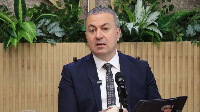 Koltukta oturan bir kişi, mikrofonun önünde konuşuyor; arka planda yeşil bitkiler var.