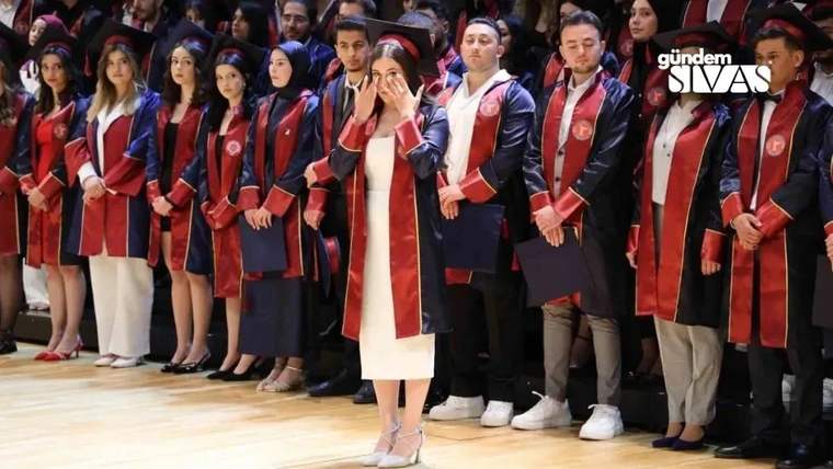 Mezuniyet cübbesi ve kepi giyen öğrenciler arasında, elini yüzüne götüren bir kadın duruyor.