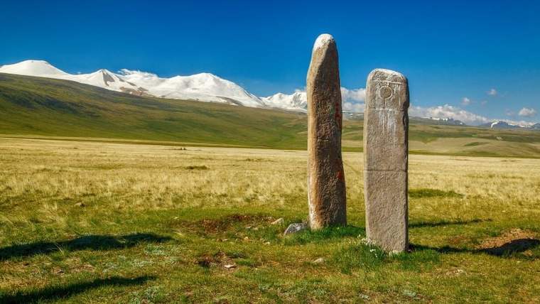 İki dikili taş, yeşil bir alanda, arka planda karla kaplı dağlar görünmekte.
