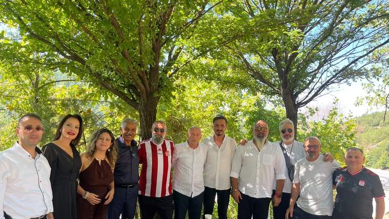 Ağaç gölgesinde bir masa çevresinde duran grup, kameraya bakarak açık havada poz veriyor.