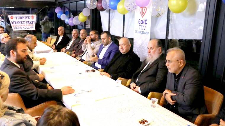 Balonlarla süslenmiş salonda, uzun masa çevresinde oturan bir grup insan toplantı yapıyor.