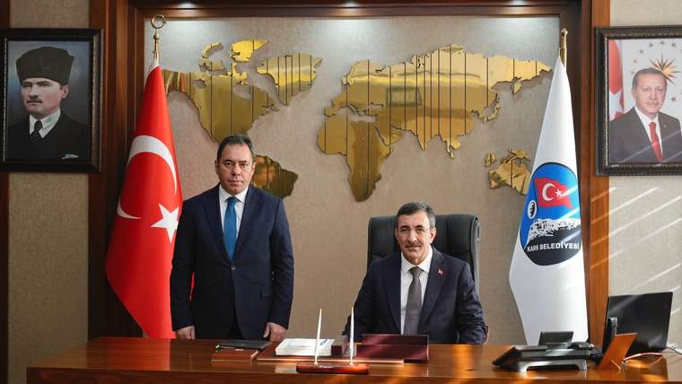 Türk bayrağı ve belediye flamaları önünde, harita bulunan duvarın önünde iki kişi masada duruyor.