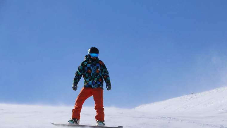 Karlı bir yamaçta, renkli mont ve kırmızı pantolon giyen kişi snowboard üzerinde duruyor.