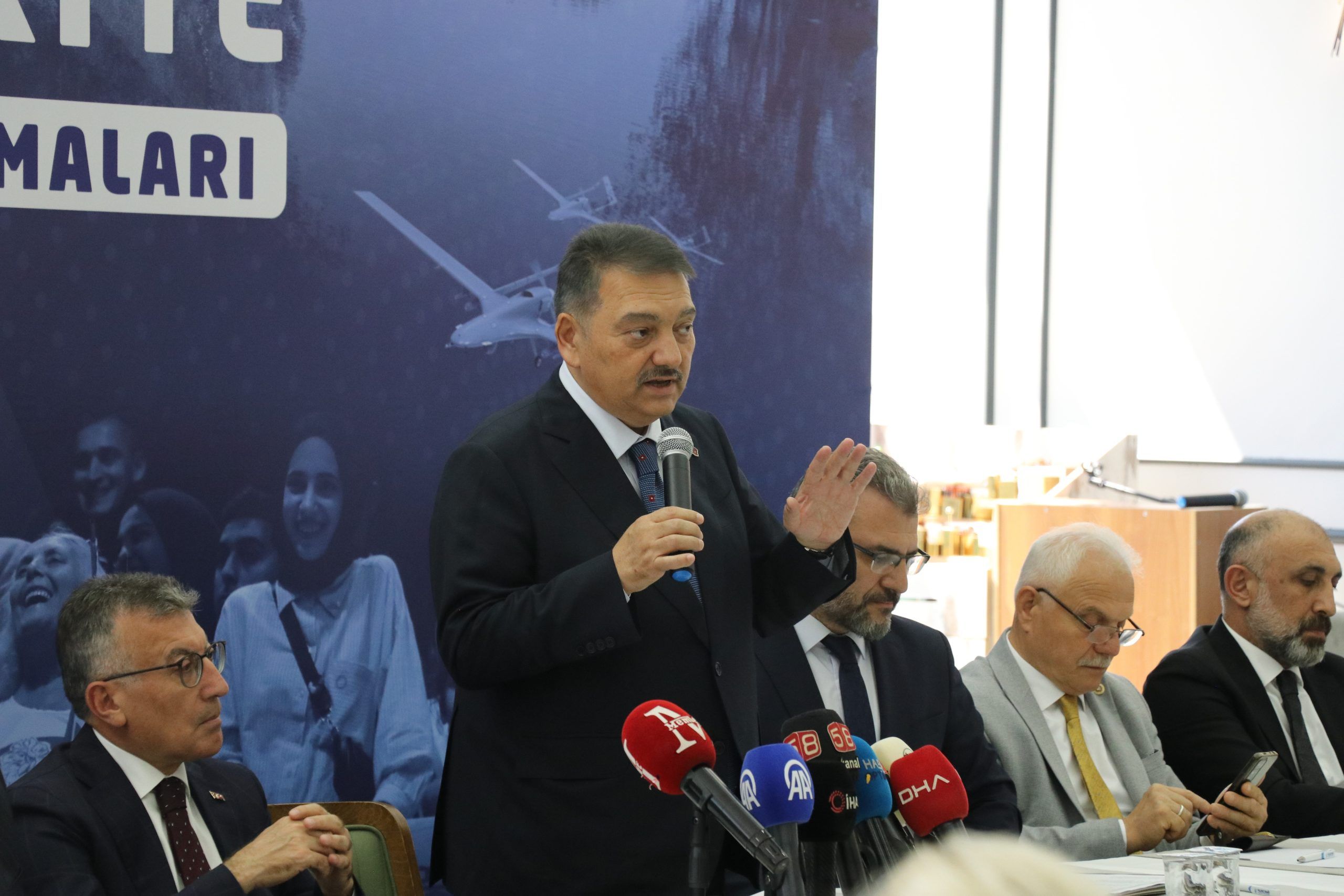 Mikrofonla konuşan bir kişi, önündeki basın mikrofonları arasında elini kaldırırken masada oturan kişiler görülüyor.