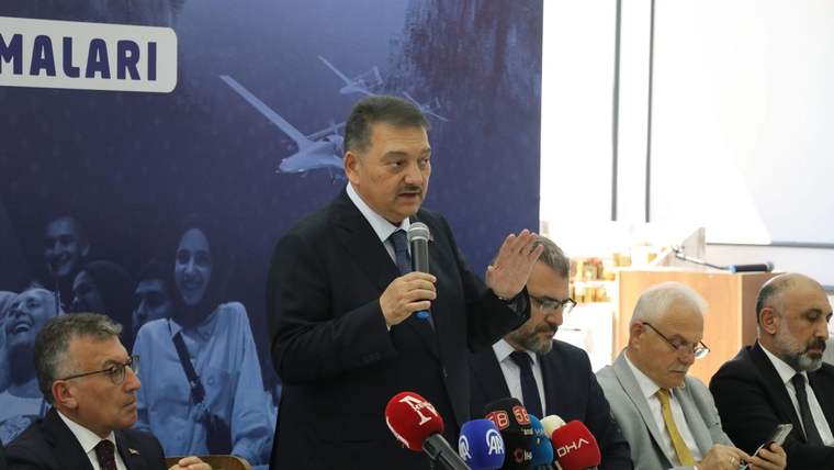 Mikrofonla konuşan bir kişi, önündeki basın mikrofonları arasında elini kaldırırken masada oturan kişiler görülüyor.