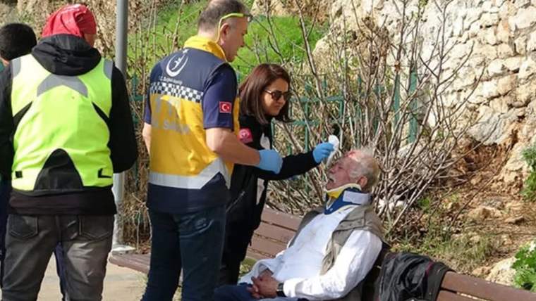 Bankta oturan yaşlı bir erkeğe, üç görevli sağlık kontrolü yapıyor; çevrede taş duvar ve bitkiler bulunuyor.