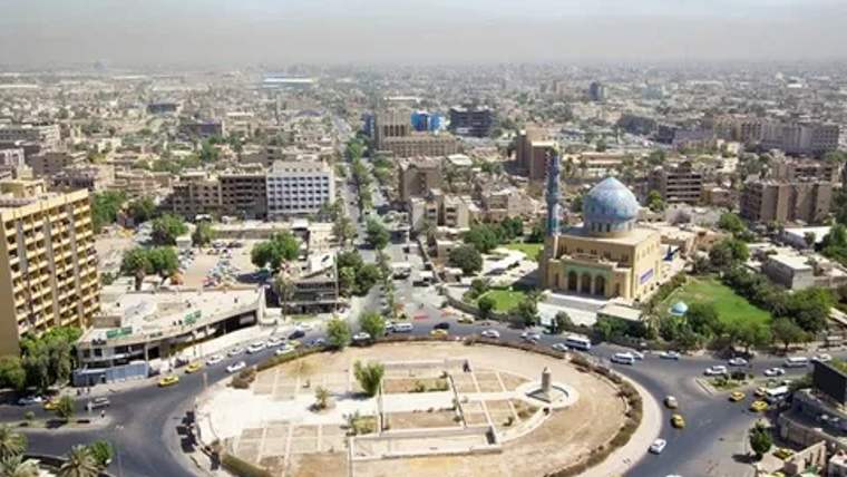 Bir şehir manzarası, ortada bir meydan ve çevresinde binalar ile araçlar görünmekte.