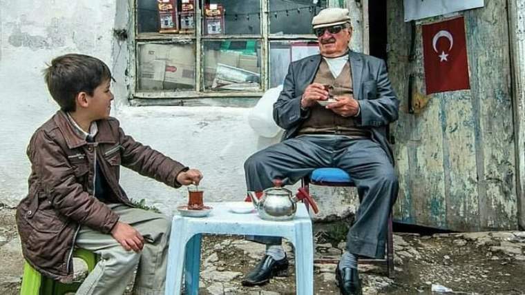 Sokak önünde oturan çocuk ve yaşlı adam, küçük masadaki çaydanlıkla birlikte görülüyor; arkada Türk bayrağı var.