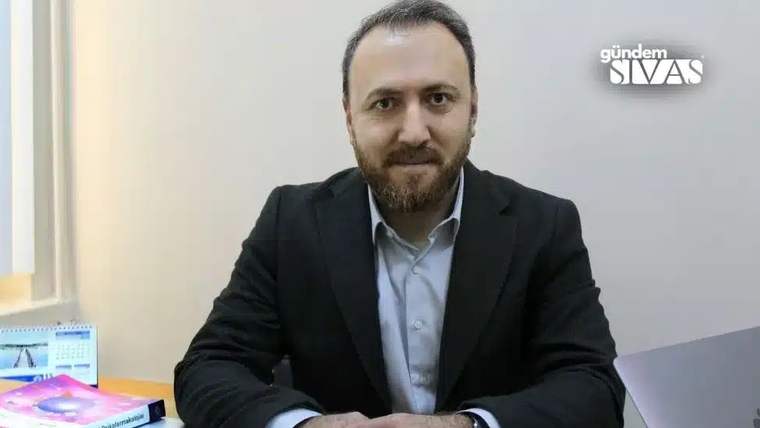 Masa başında oturan sakallı bir kişi, siyah ceket ve açık renk gömlekle kameraya bakıyor.