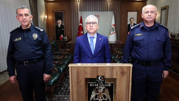 Sivas Valisi, iki güvenlik görevlisiyle birlikte bir masanın önünde duruyor.