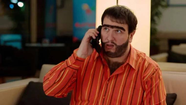 Turuncu çizgili gömlek giyen, takma kaşlı ve sakallı bir adam telefonla konuşurken görülüyor.