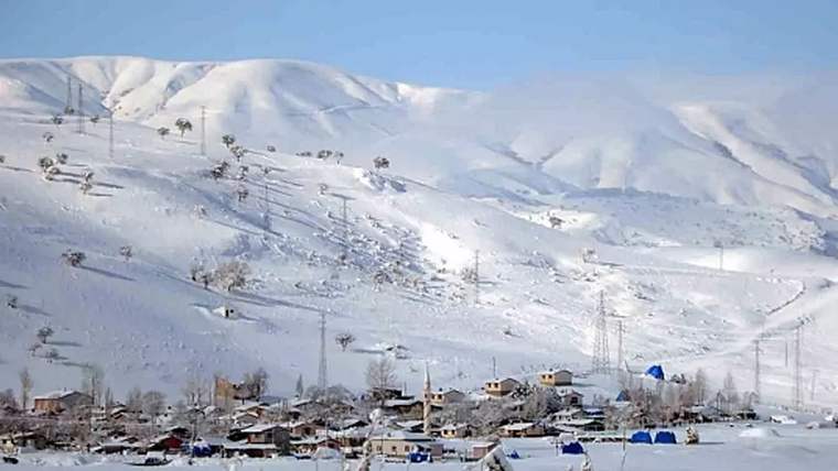 Karlarla kaplı dağların eteğinde, evlerin ve direklerin bulunduğu geniş bir kış manzarası görülüyor.