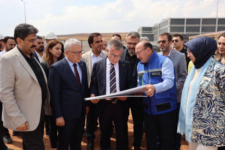 Bir grup insan, bir proje planını inceliyor; arka planda inşaat alanı görünmekte.