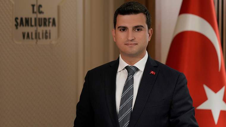 Koyu takım elbise giymiş bir kişi, arkasında Türk bayrağı ve Şanlıurfa Valiliği tabelası ile poz veriyor.