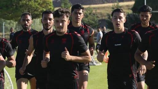 Siyah ve kırmızı spor kıyafetleri giyen futbolcular, çim sahada yan yana koşuyor.