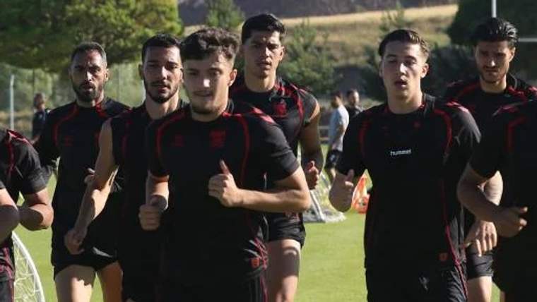 Siyah ve kırmızı spor kıyafetleri giyen futbolcular, çim sahada yan yana koşuyor.