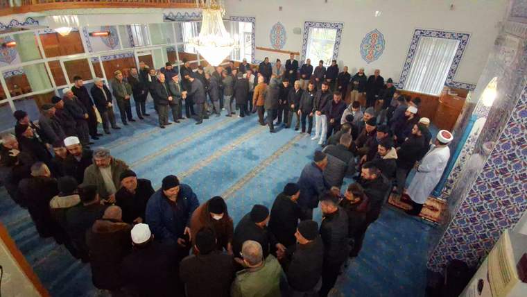 Cami içinde, mavi halılar üzerinde çok sayıda erkeğin avize ve çinilerle çevrili alanda toplandığı görülüyor.