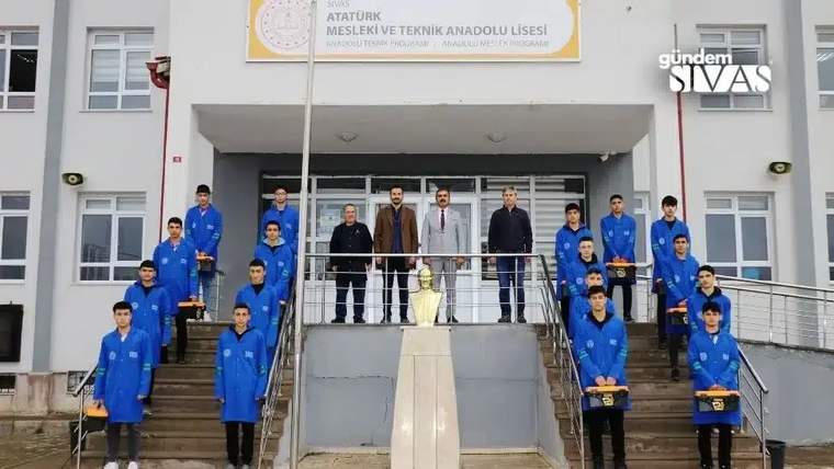 Atatürk Mesleki Teknik Anadolu Lisesi önünde mavi iş kıyafetli öğrenciler ve yetişkinler merdivenlerde duruyor.
