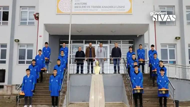 Atatürk Mesleki Teknik Anadolu Lisesi önünde mavi iş kıyafetli öğrenciler ve yetişkinler merdivenlerde duruyor.