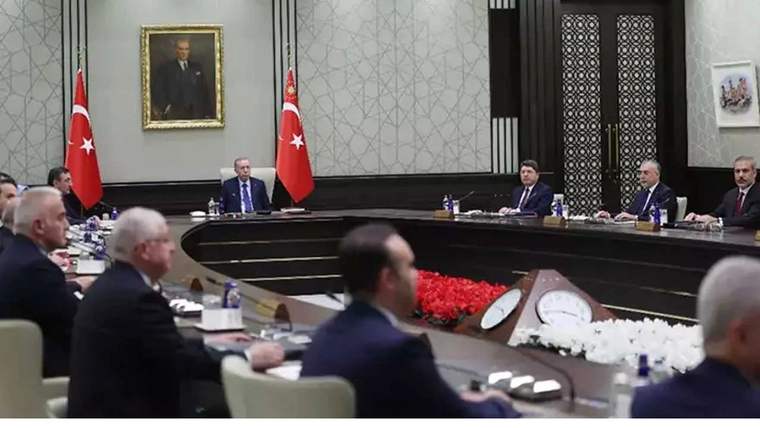 Toplantı masası çevresinde oturan kişiler, Türk bayrakları ve duvarda asılı bir portre görülüyor.