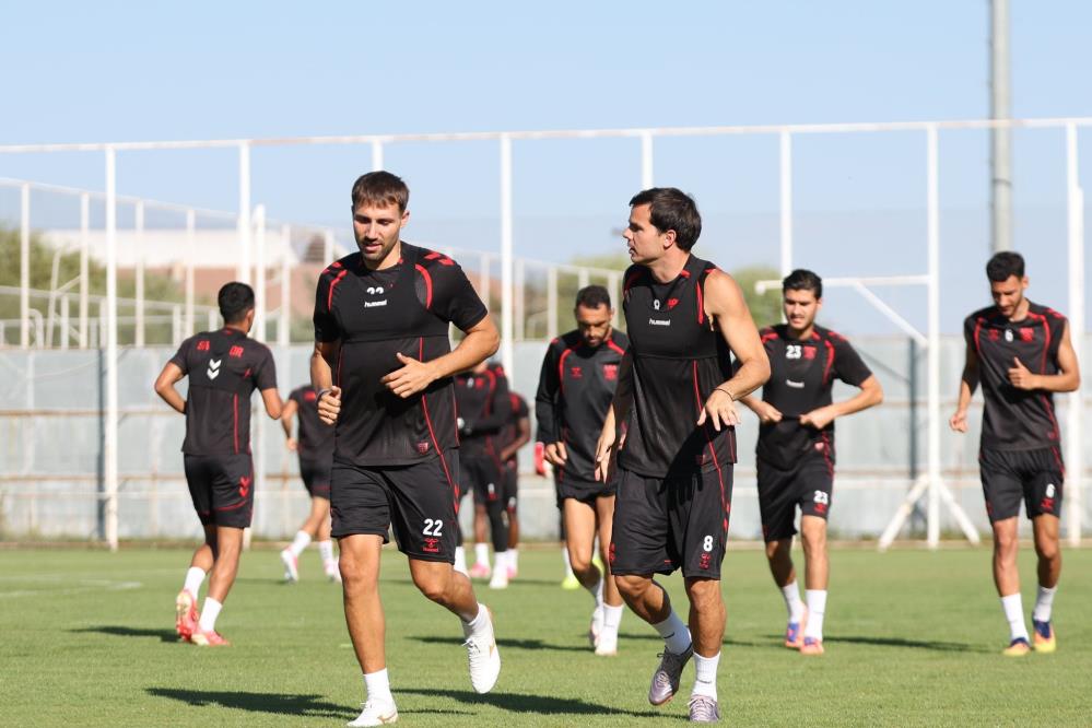 Siyah ve kırmızı antrenman formaları giyen futbolcular, çim sahada birlikte koşu yapıyor.