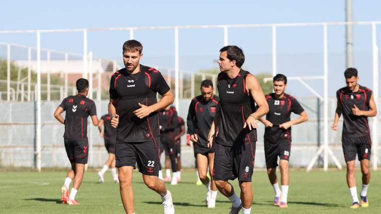 Siyah ve kırmızı antrenman formaları giyen futbolcular, çim sahada birlikte koşu yapıyor.
