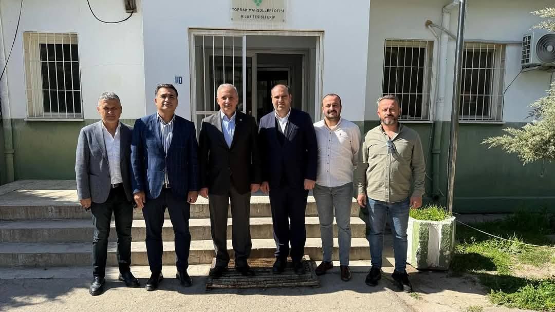 Beyaz bir binanın girişinde, takım elbiseli ve günlük giyimli altı kişi yan yana duruyor.