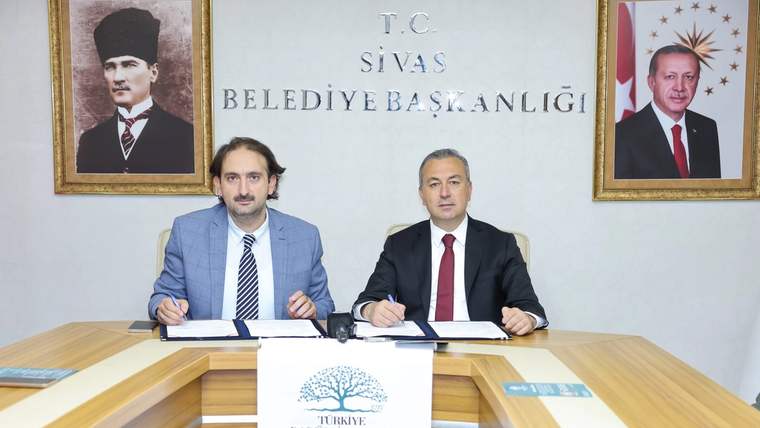 Masada belge imzalayan iki kişi, arkalarında Sivas Belediye Başkanlığı yazısı ve duvarda iki portreyle görülüyor.