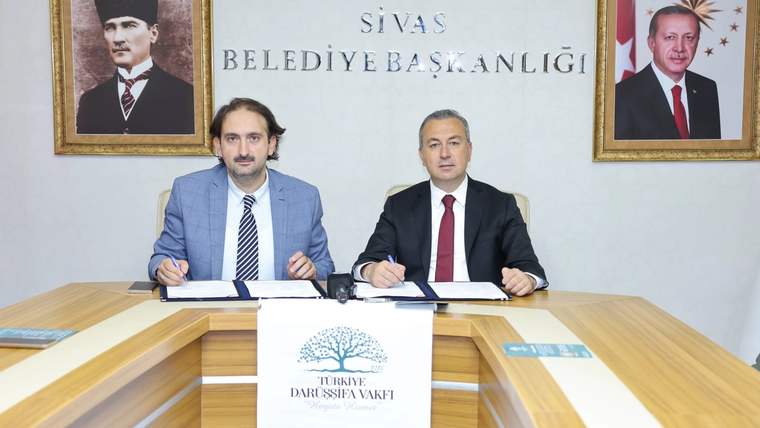 Masada belge imzalayan iki kişi, arkalarında Sivas Belediye Başkanlığı yazısı ve duvarda iki portreyle görülüyor.