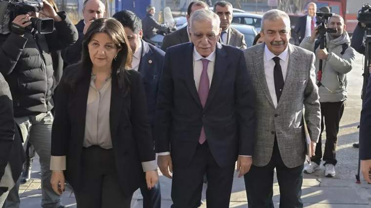 Kaldırımda yürüyen üç kişi, çevrelerinde kameralarıyla onları izleyen ve görüntüleyen insanlar bulunuyor.