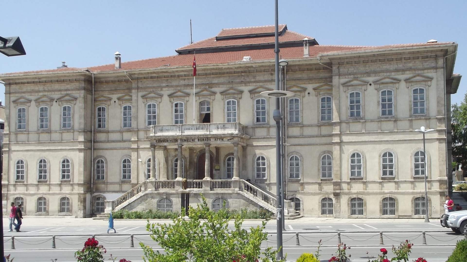 Önünde kaldırım ve çiçekler bulunan, sütunlu girişe sahip üç katlı geniş taş bina.