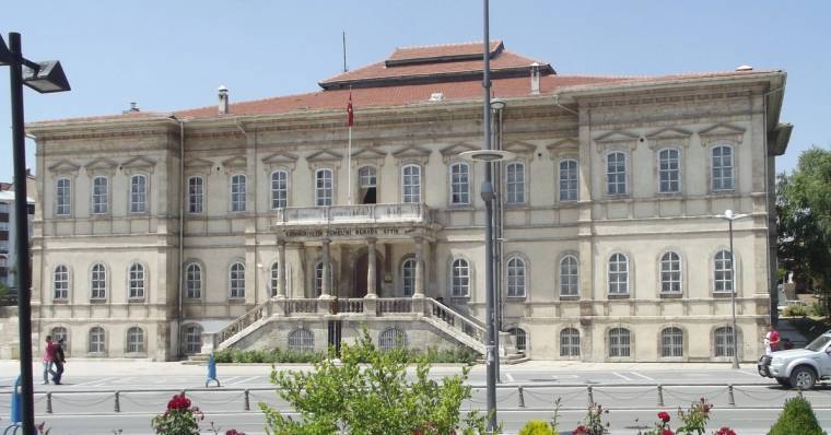 Önünde kaldırım ve çiçekler bulunan, sütunlu girişe sahip üç katlı geniş taş bina.