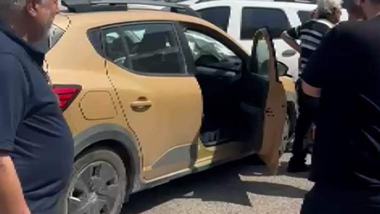 Sarı renkli bir otomobilin açık kapısı yanında üç kişi duruyor; arka planda başka araçlar ve binalar görülüyor.