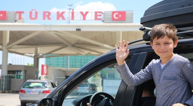 Bir çocuk, otomobilin içinde el sallayarak gülümsemekte, arka planda Türkiye sınır kapısı görünmekte.