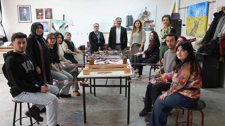 Atölye odasında bir grup insan, üzerinde renkli dokuma örnekleri bulunan masanın çevresinde oturuyor ve duruyor.