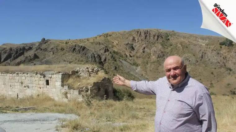 Ekose gömlekli bir adam, kayalık tepe ve taş duvar kalıntılarının önünde eliyle yön gösteriyor.