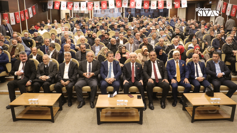 Bir toplantıda oturan kalabalık bir grup, arka planda Türk bayraklarıyla süslenmiş.