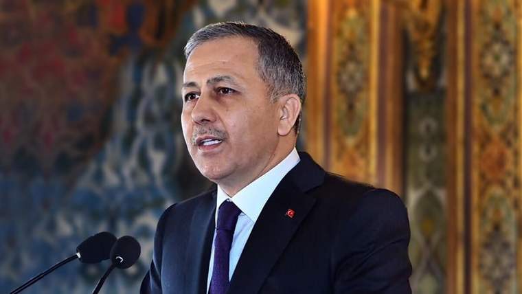 Takım elbiseli bir kişi, yakasında Türk bayrağı rozetiyle iki mikrofonun önünde konuşuyor.