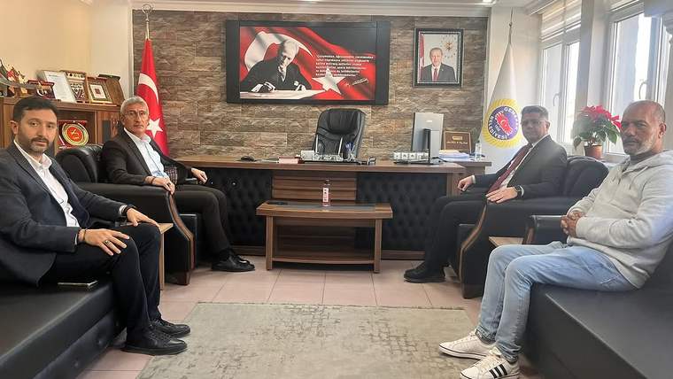 Makam odasında masa çevresinde oturan beş erkek, duvarda Türk bayrağı ve bir kurum amblemi görülüyor.