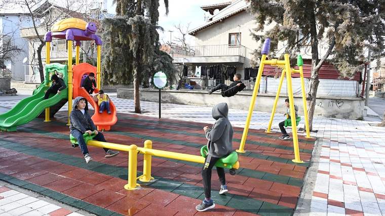 Çocuklar, renkli oyun grupları ve salıncakların bulunduğu kauçuk zeminli parkta oynuyor.