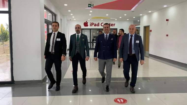 Dört takım elbiseli kişi, üzerinde “iPad Center” yazan geniş bir koridorda yürürken görülüyor.