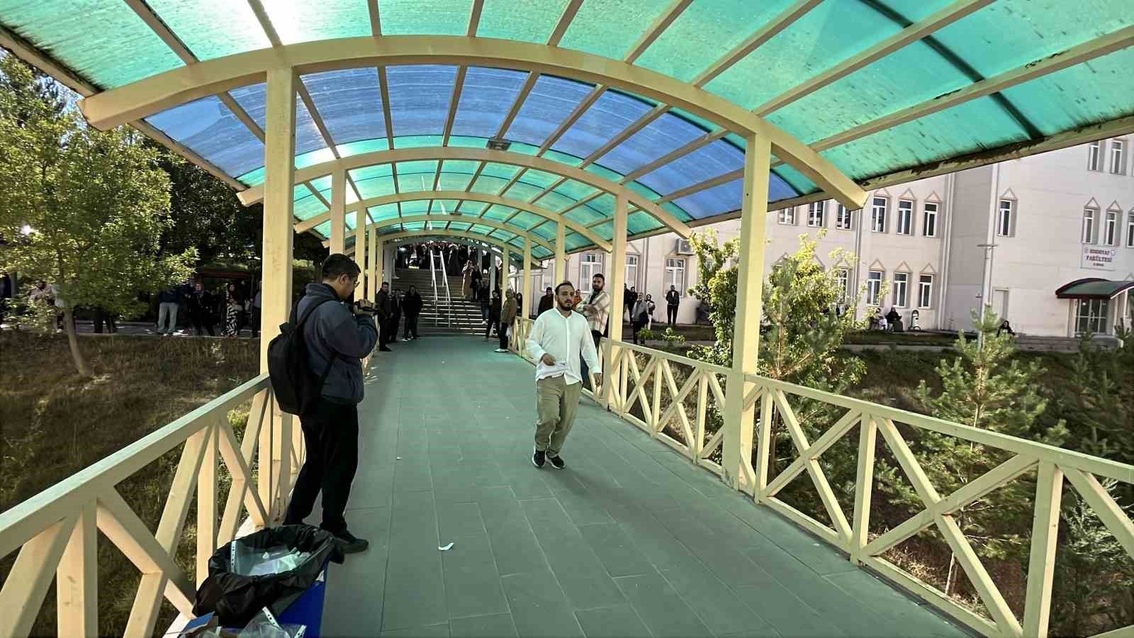 Yarı saydam çatılı kapalı yaya geçidinde iki kişi yürürken, kenarda çantalar ve ağaçlar görülüyor.
