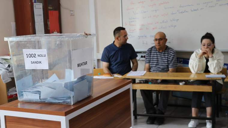 Üzerinde “1002 No’lu Sandık” ve “Muhtar” yazan oy sandığı önünde üç kişi masa başında oturuyor.