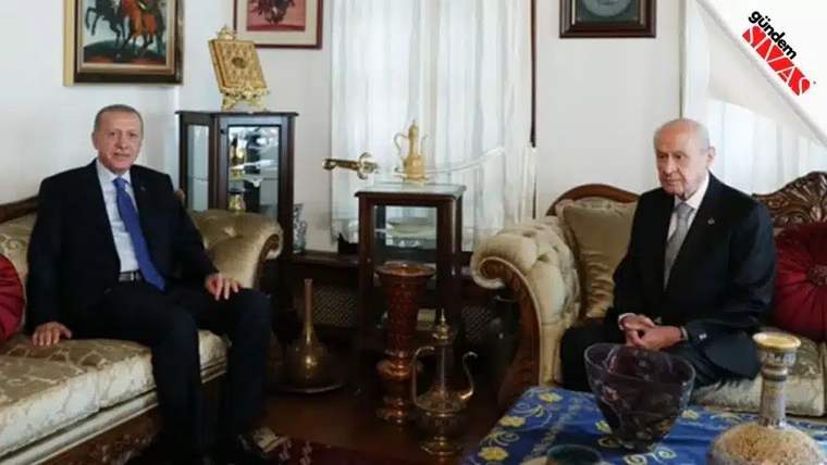 İki kişi, bir oturma odasında karşılıklı oturuyor; arka planda vitrin ve dekoratif eşyalar var.