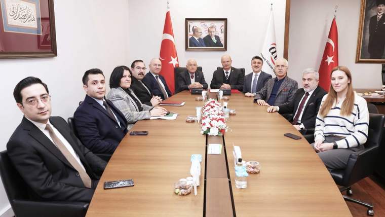 Toplantı masası çevresinde oturan bir grup, Türk bayrakları ve çiçek aranjmanı önünde poz veriyor.