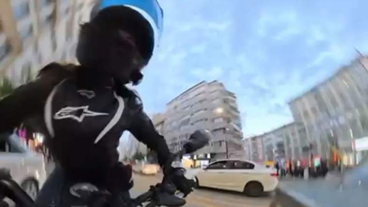 Kasklı motosiklet sürücüsü, şehir içindeki yolda ilerlerken yanında beyaz bir otomobil görülüyor.