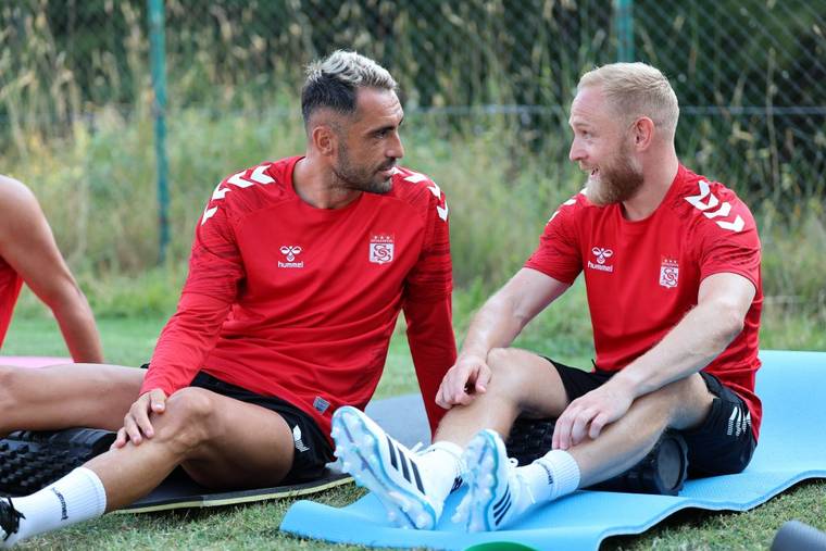 Kırmızı forma giymiş iki futbolcu, antrenman alanında oturarak sohbet ediyor.