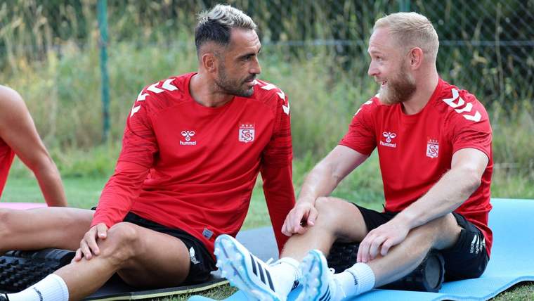 Kırmızı forma giymiş iki futbolcu, antrenman alanında oturarak sohbet ediyor.
