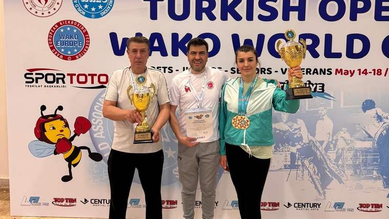 Üç kişi, madalyalar ve kupalarla birlikte poz veriyor; arka planda turnuva afişi var.