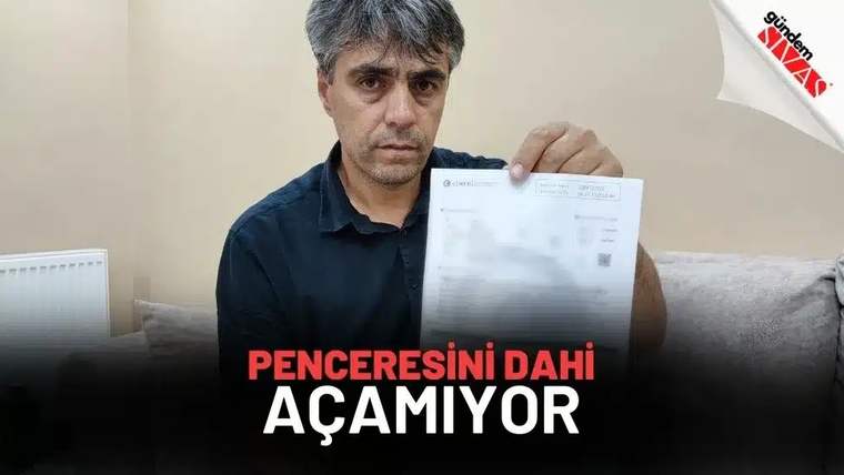 İç mekânda bir erkek, üzerinde yazılar bulunan bir belgeyi kameraya doğru tutuyor; arka planda koltuk görülüyor.